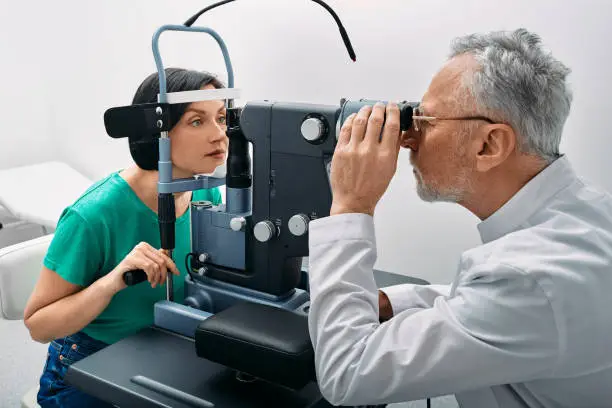 Woman while eye exam with binocular slit-lamp at ophthalmology clinic with experienced gray-haired optometrist