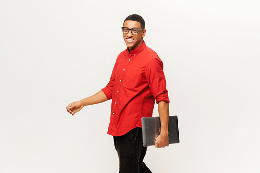 Cheerful handsome smiling African-American young man in red shirt carrying laptop and walking isolated on white. Friendly well-looking multiracial student, freelancer in glasses