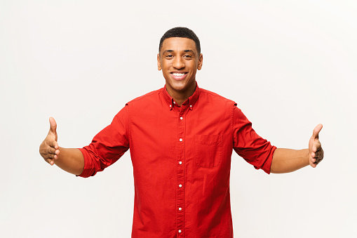 Cheerful Aferican-American young man holding two hands in front of him and shows the wide size, latin guy in red shirt advertising novelty, presenting some product isolated on white background