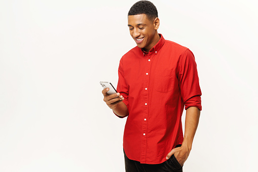 Smiling optimistic African-American young man in red casual shirt using smartphone isolated on white, multiracial freelancer or student gay chatting online, texting, messaging, web surfing, copy space