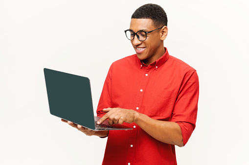 Cheerful handsome smiling African-American young man in red shirt using laptop isolated on white. Friendly smart multiracial student, freelancer in glasses typing, using computer for remote work