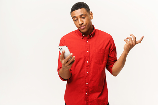 Confused young African-American short-cutted guy in red shirt holds smartphone and shrugs shoulders isolated on white background, disappointed multiracial man looks at phone screen