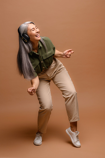Happy crazy elderly mature woman in headphones signs isolated on brown. Overjoyed senior grey-haired female laughing and singing