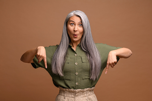Cheerful positive woman pointing fingers down showing space for your advertisement, looking at camera with surprised face. Indoor studio shot isolated on brown background