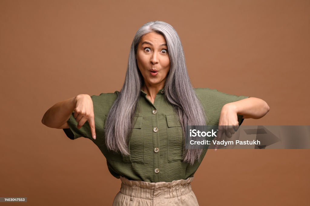 Cheerful positive woman pointing fingers down showing space for your advertisement, looking at camera with surprised face. Indoor studio shot isolated on brown Cheerful positive woman pointing fingers down showing space for your advertisement, looking at camera with surprised face. Indoor studio shot isolated on brown background Pointing Stock Photo