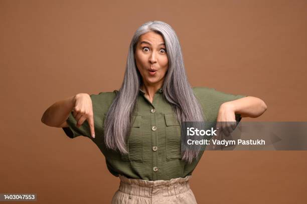 Cheerful Positive Woman Pointing Fingers Down Showing Space For Your Advertisement Looking At Camera With Surprised Face Indoor Studio Shot Isolated On Brown Stock Photo - Download Image Now