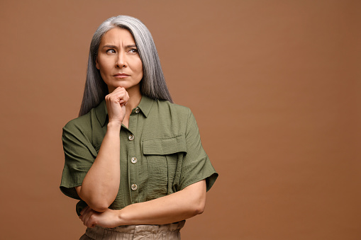 Smart and thoughtful mature woman holding her chin and pondering idea, making difficult decision, looking uncertain doubtful. Indoor studio shot isolated on beige background
