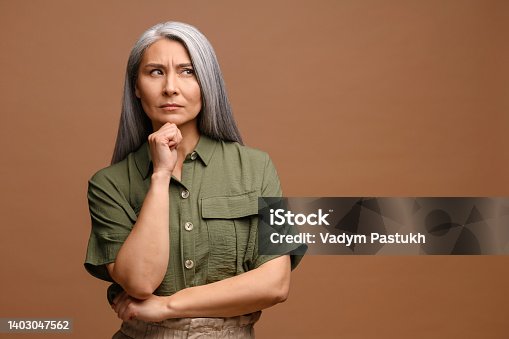istock Smart and thoughtful mature woman holding her chin and pondering idea, making difficult decision, looking uncertain doubtful. Indoor studio shot isolated on beige 1403047562