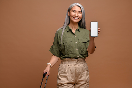 Cheerful senior asian woman in casual wear stands with suitcase and showing phone with empty screen, isolated on brown, woman advertising mobile app for travel