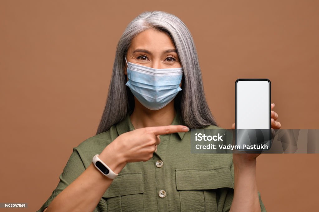 World pandemic, epidemic. Mature woman in medical mask with a smartphone in her hand, she points a finger at the screen World pandemic, epidemic. Mature woman in medical mask with a smartphone in her hand, she points a finger at the empty screen, mockup concept. Isolated on brown background Epidemic Stock Photo