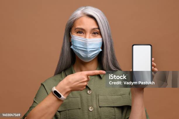 World Pandemic Epidemic Mature Woman In Medical Mask With A Smartphone In Her Hand She Points A Finger At The Screen Stock Photo - Download Image Now
