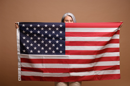 Happy and excited asian woman hiding behind the American flag while standing over brown isolated background, peeking out from behind the usa flag. Patriotism concept