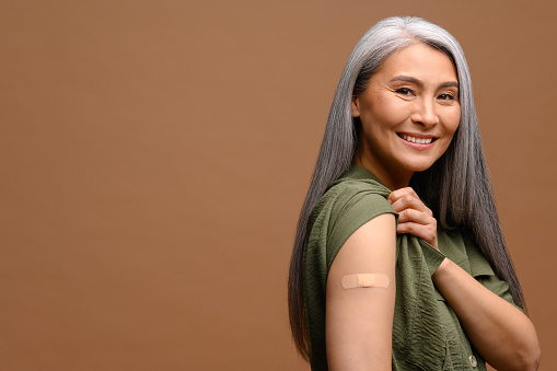 Smiling senior grey-haired woman with a medical patch after vaccination isolated on brown background, protecting hand with bandage after injection. Healthcare and medicine concept
