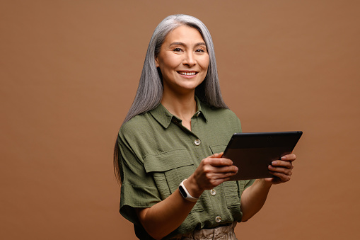 Modern elderly senior woman using digital tablet isolated on brown. Portrait of mature female office employee using online technology for doing business