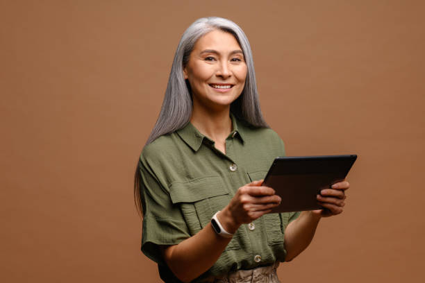 Modern elderly senior woman using digital tablet isolated on brown. Portrait of mature female office employee using online technology for doing business Modern elderly senior woman using digital tablet isolated on brown. Portrait of mature female office employee using online technology for doing business, computer app for accounting old woman smiling tablet stock pictures, royalty-free photos & images