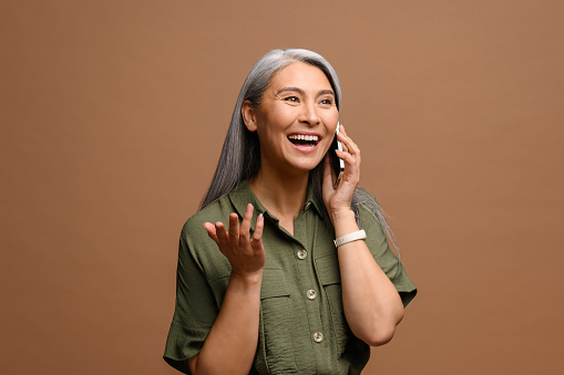 Mature lady has pleasant phone conversation. Cheerful smiling senior grey-haired business woman talking on smartphone standing isolated on brown background