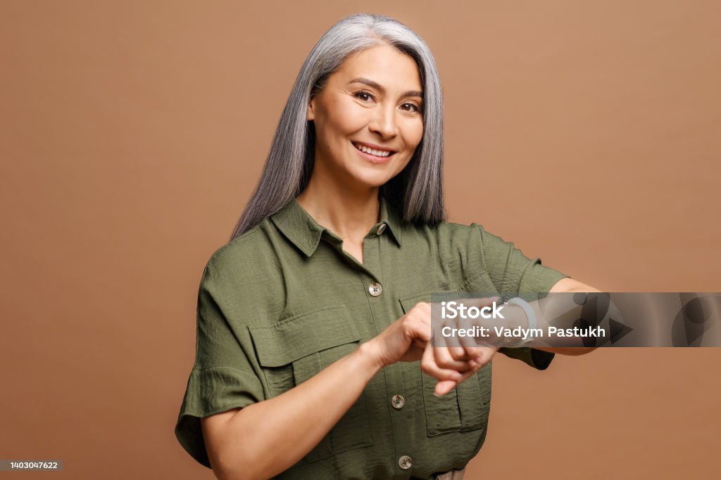Mature beautiful asian woman with grey hair wearing casual clothes In hurry pointing to watch time, impatience, talking about deadline Mature beautiful asian woman with grey hair wearing casual clothes In hurry pointing to watch time, impatience, talking about deadline delay Checking the Time Stock Photo