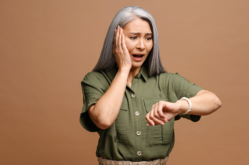 Portrait of shocked mature woman holding hand with wrist watch and looking at it with surprised expression isolated on a brown background