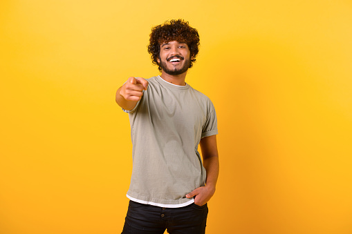 Smiling young indian man with curly hairstyle pointing finger at you standing isolated on yellow background, cheerful teenage guy indicating at the camera