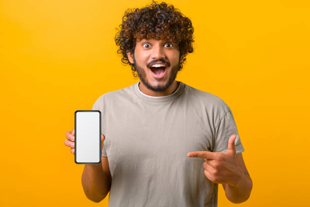 istock Excited young curly Indian guy showing smartphone and points finger at empty phone screen 1403048593