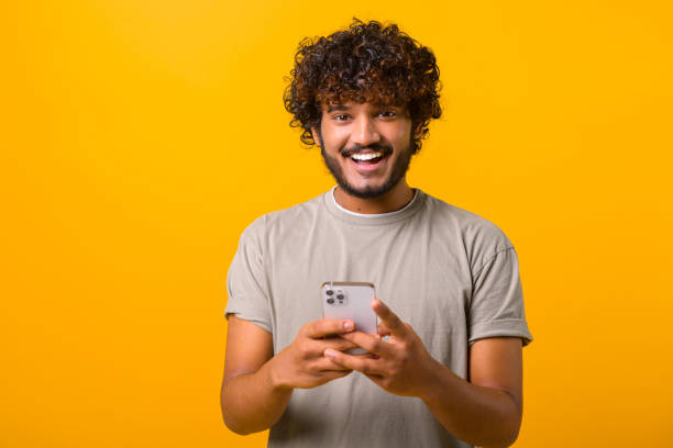 istock Ecstatic curly young Indian man standing isolated on yellow and using trendy smartphone 1403048609