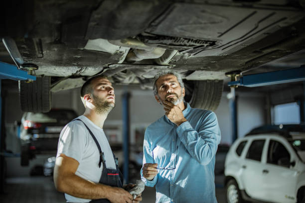 Mid adult customer and auto repairman analyzing chassis in a workshop. Male manager and auto mechanic examining undercarriage of a car in a workshop. chassis stock pictures, royalty-free photos & images