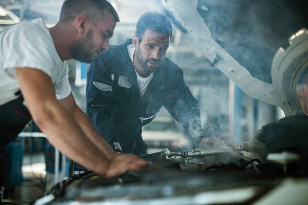 Auto mechanics working on overheated car engine in a workshop. Auto mechanics cooperating while examining overheated car engine in auto repair shop. Focus is on man in blue uniform. overheated stock pictures, royalty-free photos & images