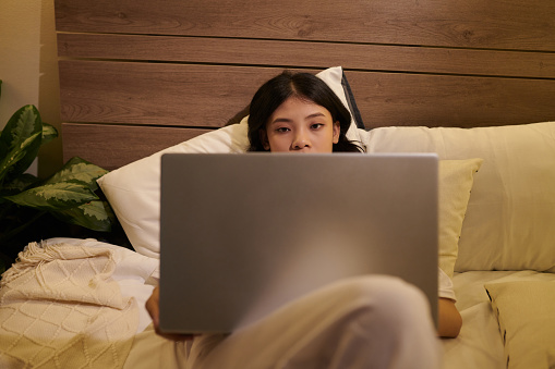 Tired young woman lying in bed with opened laptop, reading article or watching video