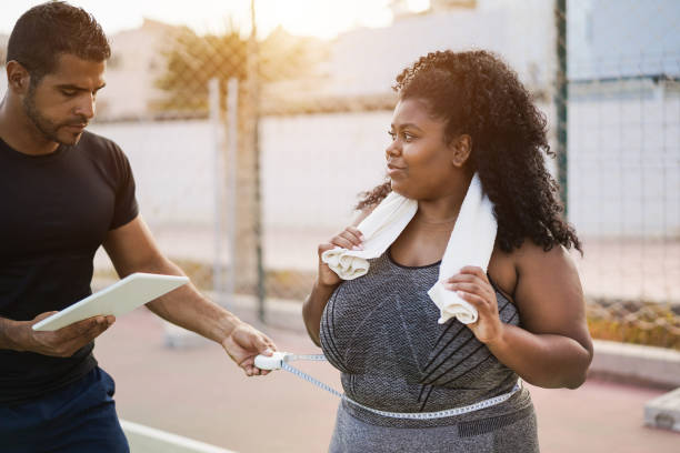Curvy woman with her personal trainer measuring body waist outdoor - Focus on african girl face Curvy woman with her personal trainer measuring body waist outdoor - Focus on african girl face waist trainer for men stock pictures, royalty-free photos & images