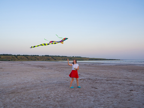 Woman controls colored kite at sunset on the beach. Concept of spending great time at summer. Relax. Bird soars in the sky. Flying kite with an hangs in air. Tail of kite sways in wind. Freedom