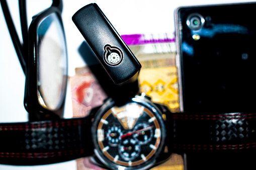 this is an hd image of an Black Zippo Lighter In Focus And An Watch An Smartphone An Eyeglass And Some Currency In Background image was shot inside an room on an table in  normal room light .