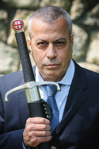 Portrait of handsome businessman model man in a modern casual suit with a large medieval sword. Modern fashion street style concept. Elegant urban warrior. Corporate and money security