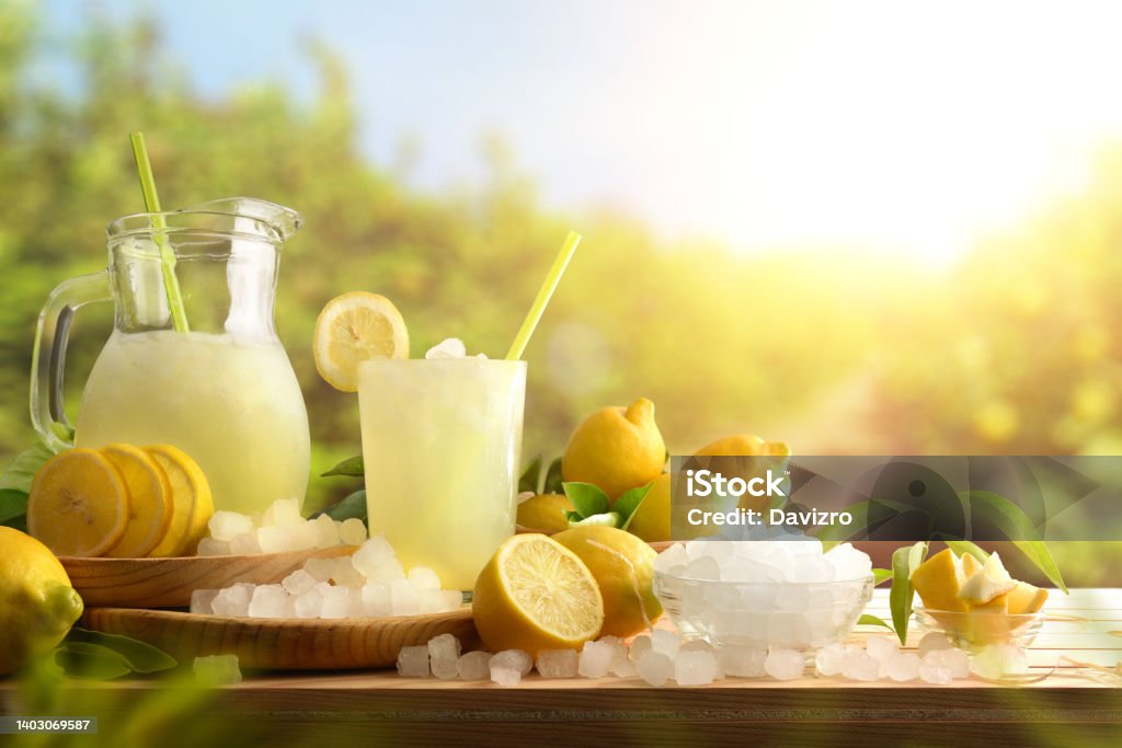 Lemonade with ice on table with lemon trees in background Lemonade with ice in a pitcher and glass on a wooden table with fruit and crushed ice outside with a lemon field in the background on a sunny day. Front view. Horizontal composition. Lemonade Stock Photo