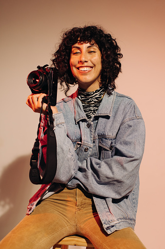 Cheerful female photographer smiling at the camera while holding a digital camera. Happy freelance photographer sitting on a chair against a studio background.