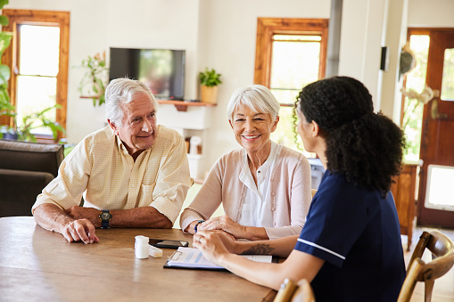 Senior couple smiling while talking with a female healthcare worker at a table in their lounge during a home visit