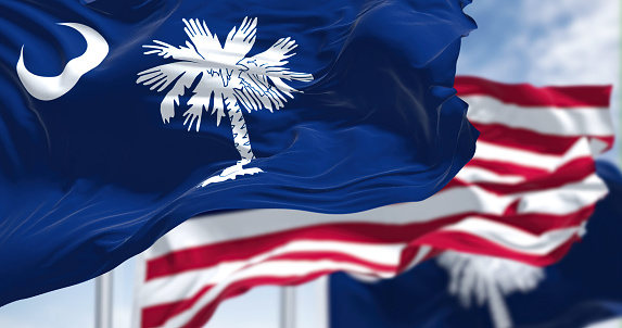 The South Carolina state flag waving along with the national flag of the United States of America. South Carolina is a state in the coastal Southeastern region of the United States. White palmetto tree on an indigo field. The canton contains a white crescent.