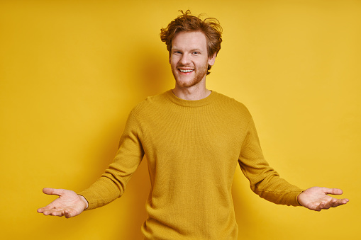 Cheerful redhead man gesturing welcome sign while standing against yellow background