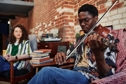 Young man in eyeglases and casualwear sitting in armchair in living room of loft apartment at leisure and playing violin for his girlfriend