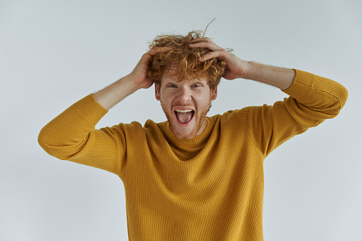 Happy young redhead man holding hands in hair while standing against wall