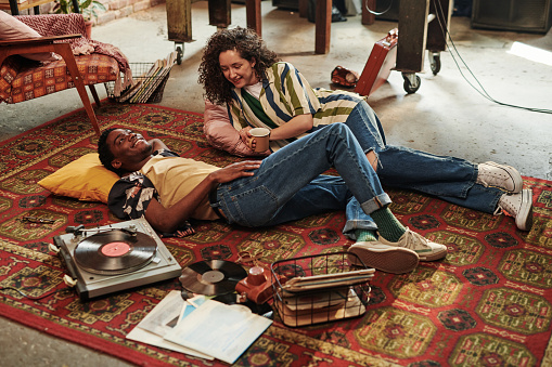 Happy young couple in stylish casualwear listening to vynil disks and talking while relaxing on red carpet on the floor of living room