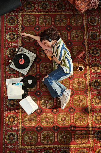 Above view of young woman in jeans and shirt looking at record player with vynil disk while listening to music in headphones on the floor