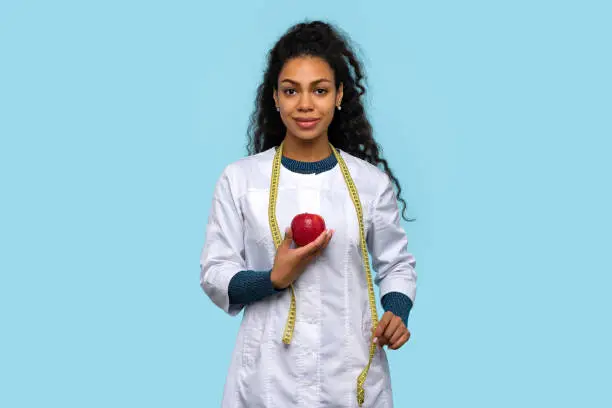 African American Woman Doctor Nutritionist wears White Coat. Good Looking Dietitian in Medical Uniform with Red Apple and Measuring Tape on Blue Studio Wall. Healthcare Worker Female Medical Employee