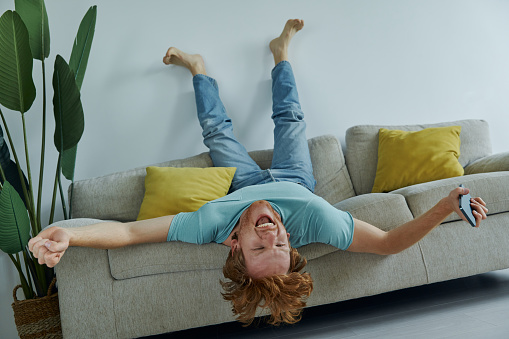 Excited redhead man holding smart phone and gesturing while relaxing on the couch at home