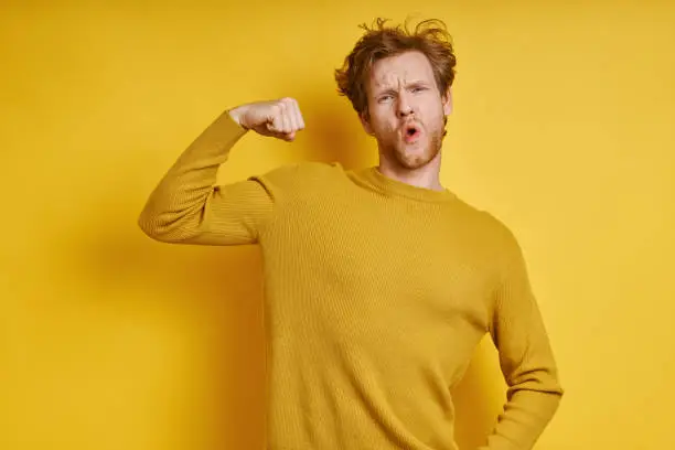Playful redhead man showing his bicep while standing against yellow background
