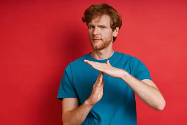 Handsome redhead man gesturing time out while standing against red background