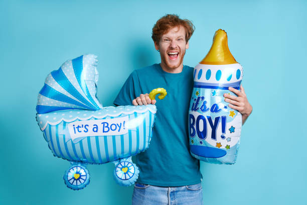 Happy redhead man holding baby stroller and milk bottle while standing against blue background