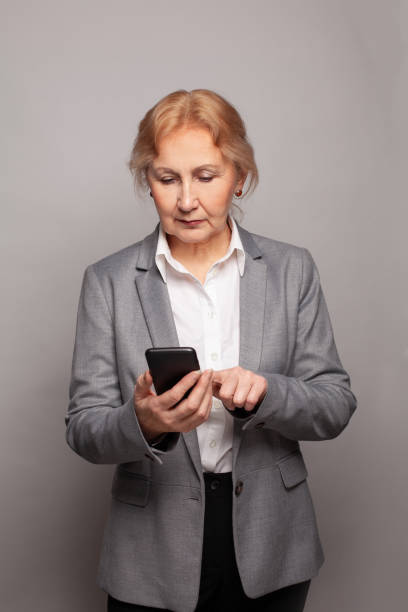 istock Serious senior woman texting with smartphone on gray studio wall background 1403087424