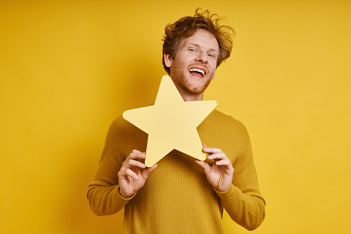 Handsome redhead man holding star shape banner and smiling against yellow background