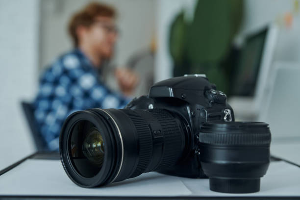 istock Close-up of digital camera and young man using computer on background 1403087819