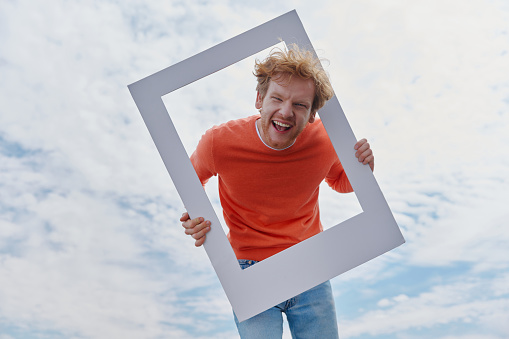 Playful young redhead man looking through a picture frame and smiling while standing outdoors